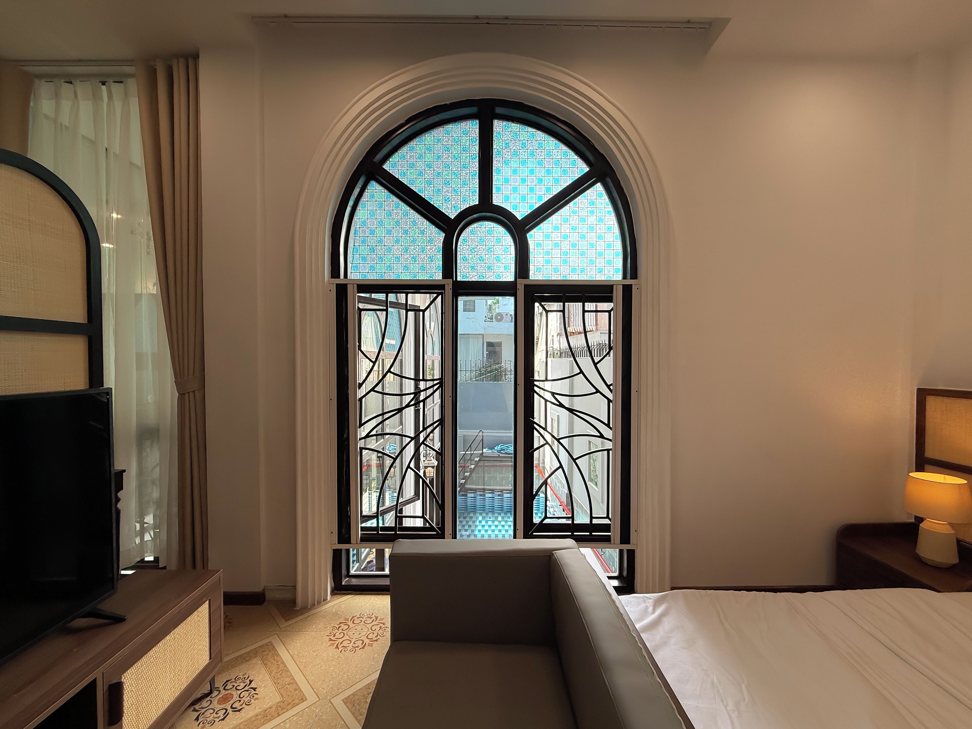 Habitación de hotel con un amplio ventanal de arco, una cama, una mesita de noche con lámpara y un televisor sobre un mueble.