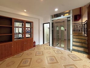 A hotel lobby with a wooden cabinet, a glass elevator, and a staircase. - Maison - Product by Qunh Khang (Tân Sơn Hòa)