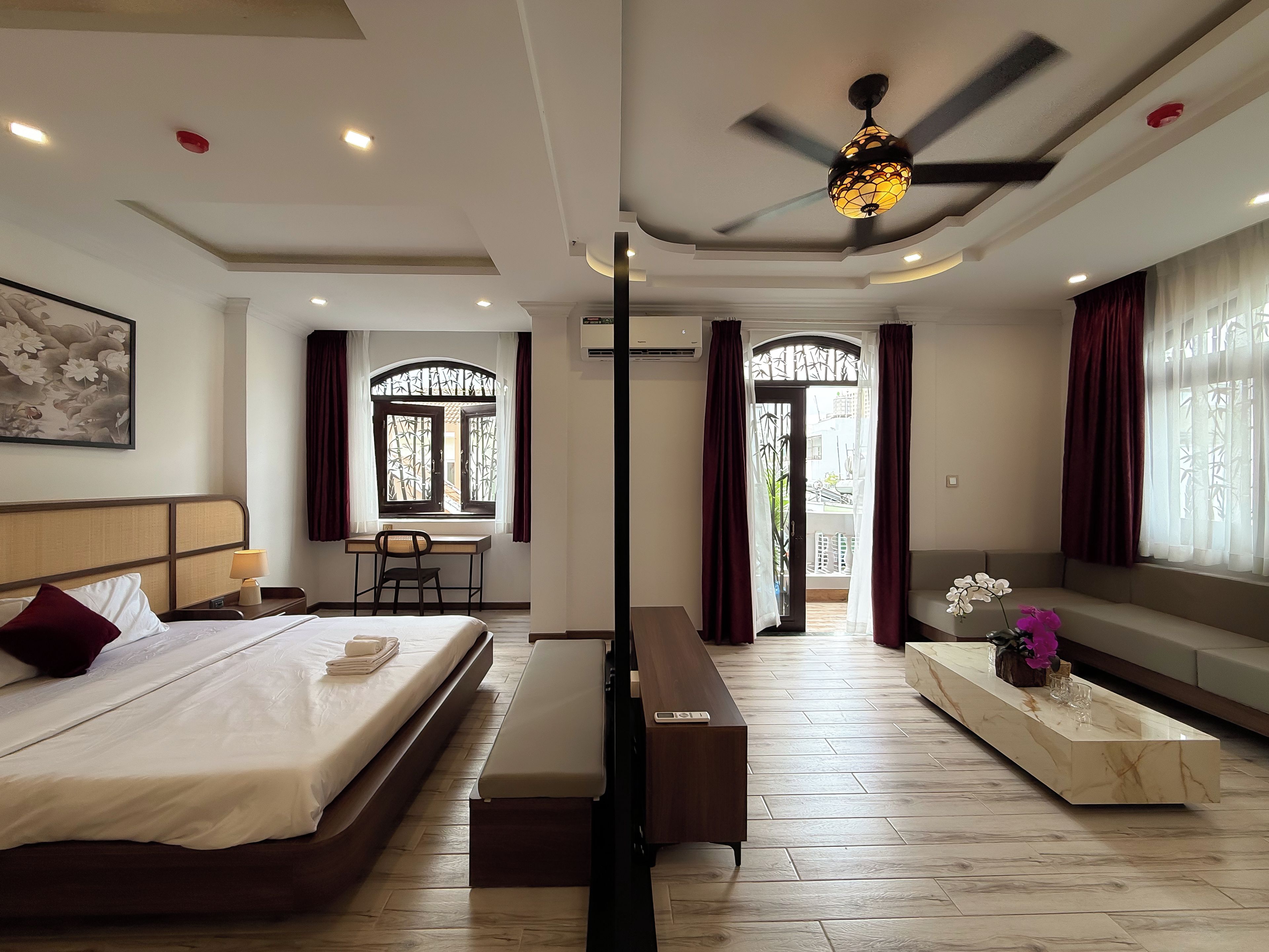 A hotel room with a bed, desk, bench, and a view through a window with curtains.