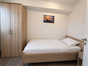 En enkeltseng med træstel, hvidt sengetøj, et træskab og et lille natbord med en lampe.