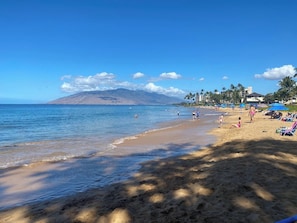 Ein Strand mit blauem Himmel, einem Berg im Hintergrund und Menschen, die das Wasser genießen.