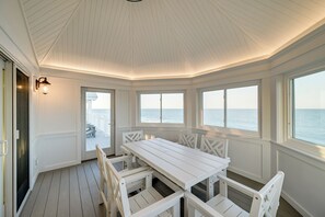 Ein helles Interieur mit Holzterrasse, Tisch und Stühlen sowie großen Fenstern mit Meerblick.