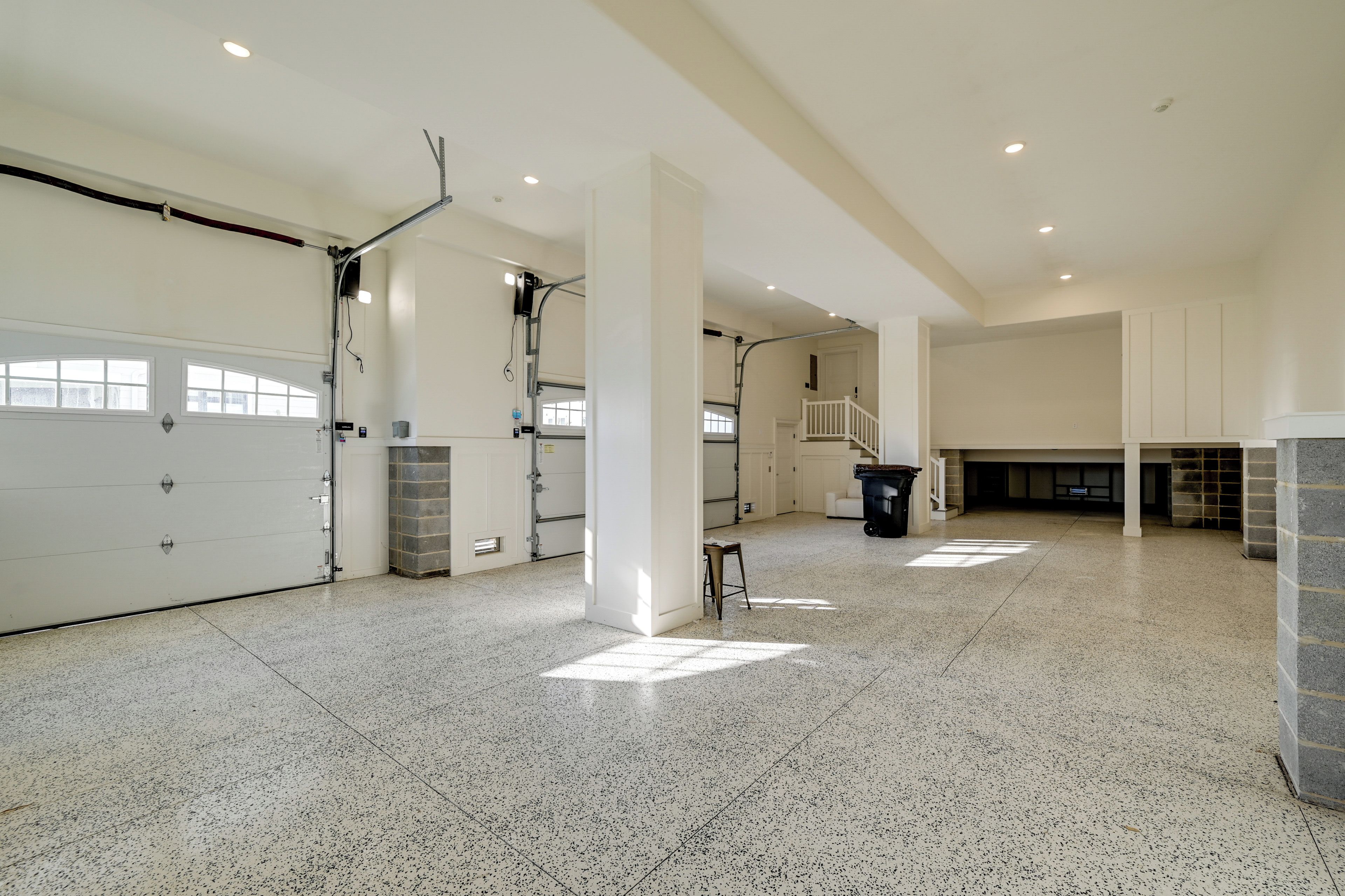 A spacious garage with a large white garage door, a staircase leading to an upper level, and a trash bin.