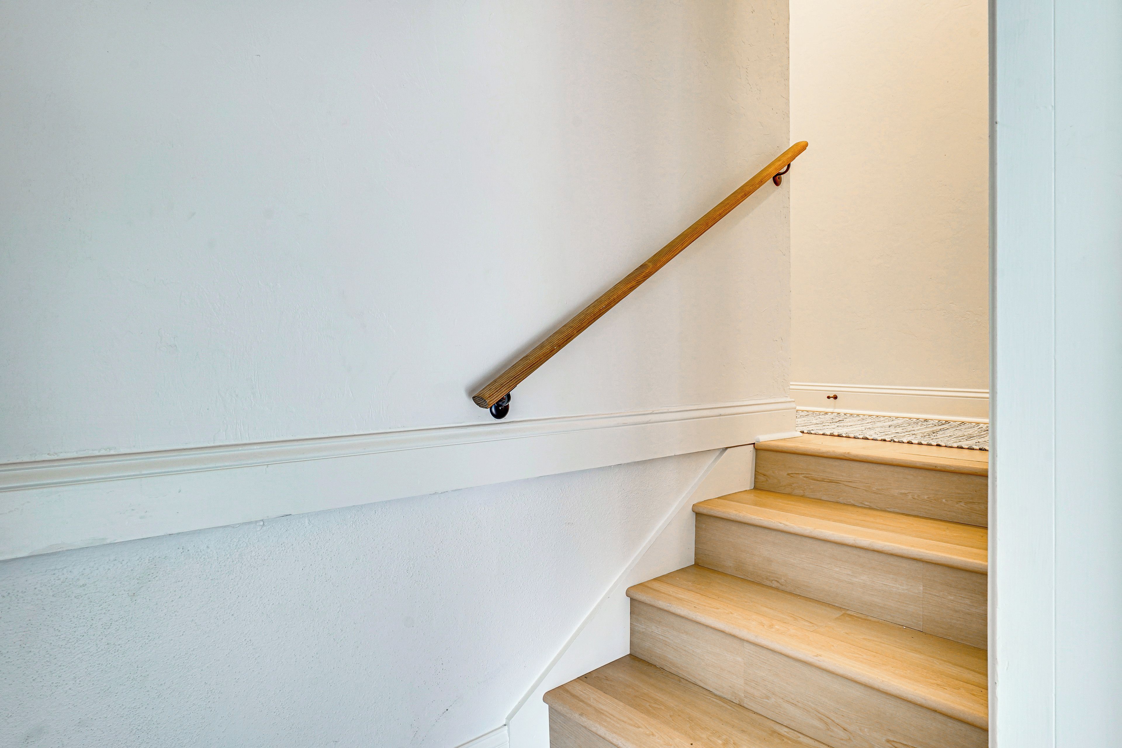 A wooden staircase with a handrail, white walls, and a textured floor.
