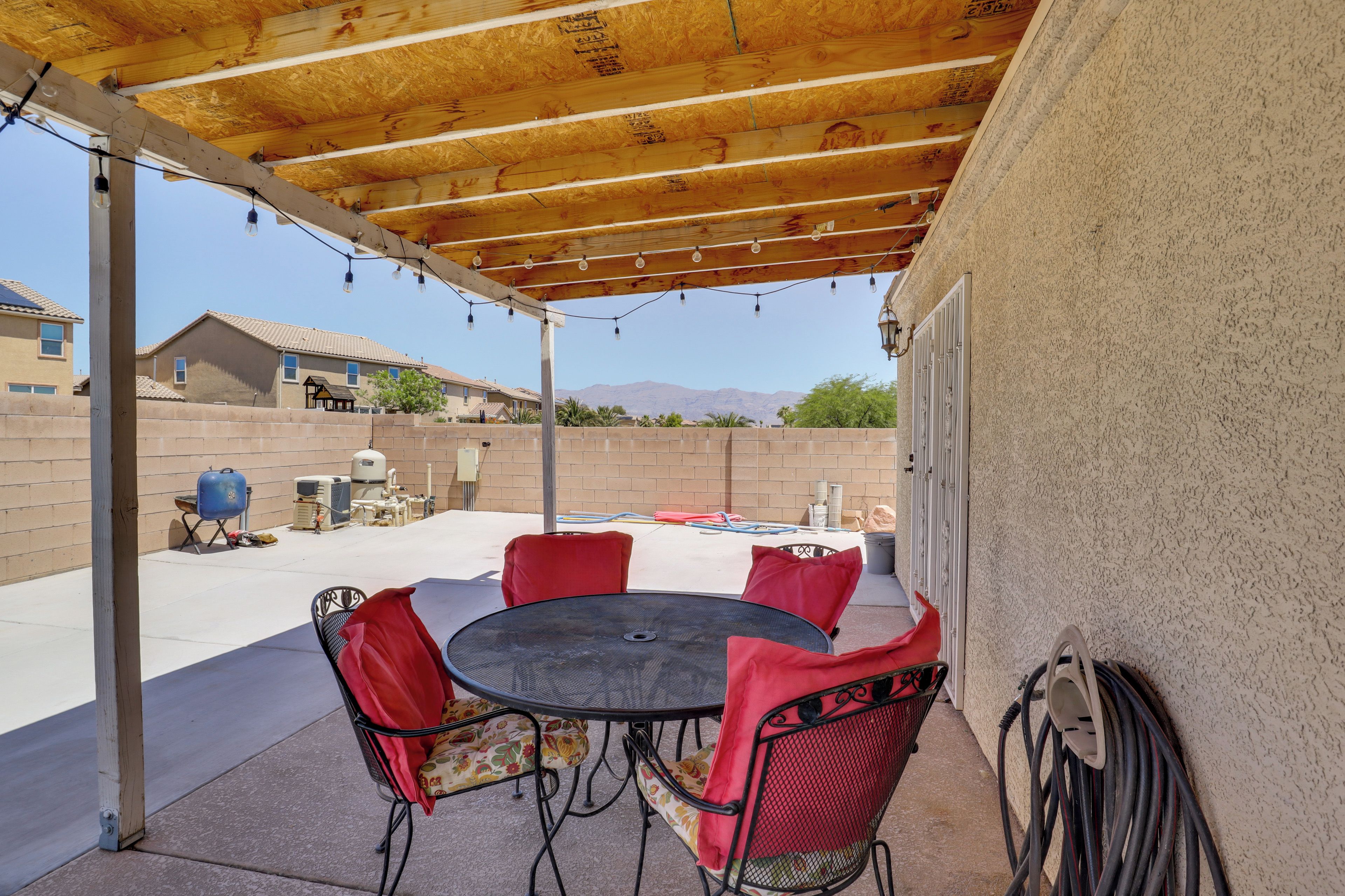 An outdoor patio with a round table and chairs, a pergola overhead, and a view of a residential area with houses and mountains in the background.