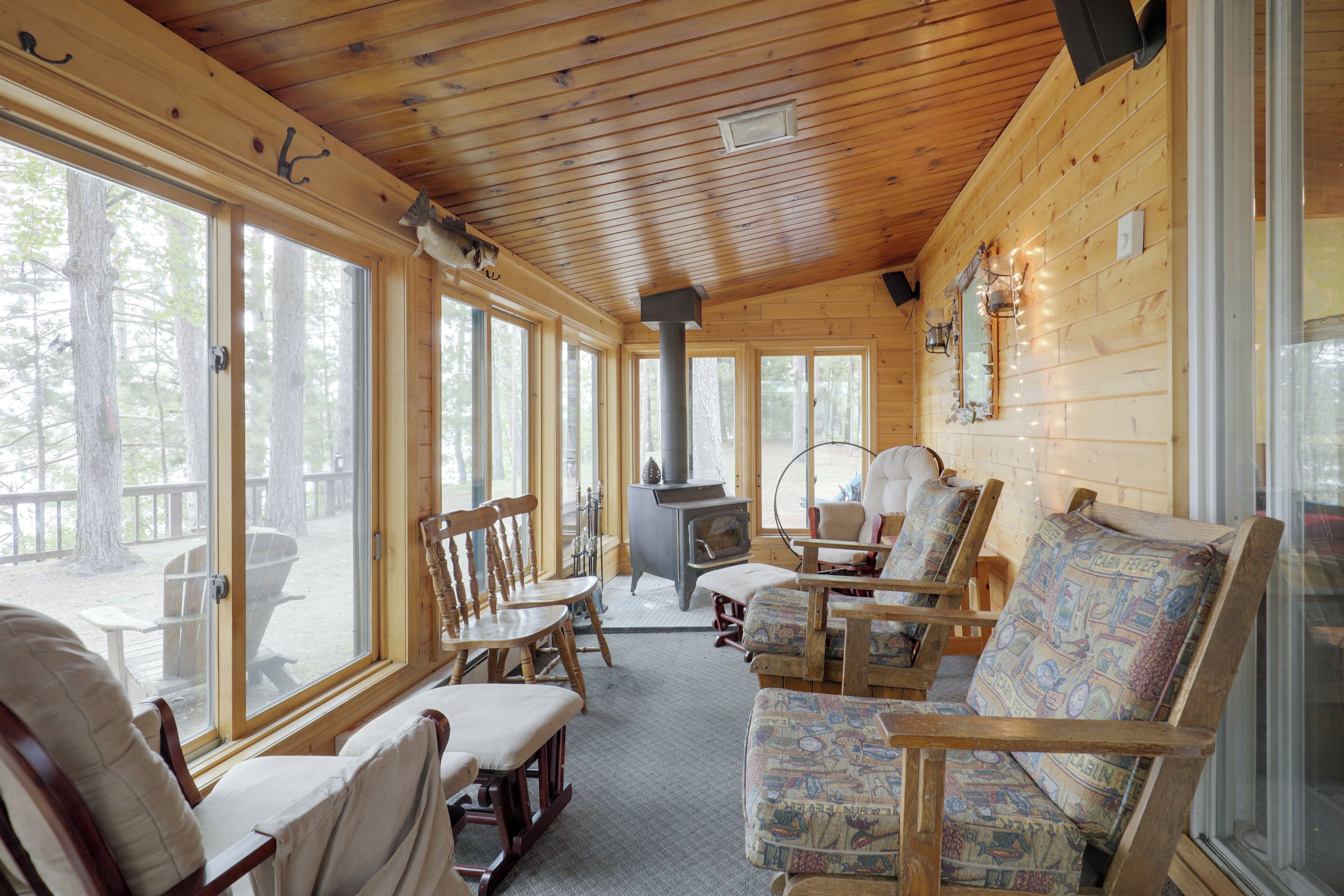 A cabin interior with wooden walls, large windows, a fireplace, and several chairs.