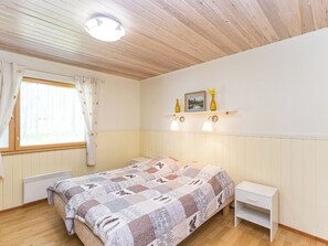 Une chambre à coucher comprenant un lit, une table de chevet, une fenêtre avec des rideaux, une étagère fixée au mur avec deux lampes, et un luminaire au plafond.