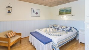 A bedroom with a wooden ceiling, a bed with a patterned bedspread, a nightstand, a chair, and wall-mounted artwork. - Hakojärventie 3b by Interhome (Kuusamo)