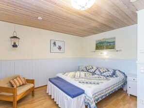 Un dormitorio con techo de madera, una cama con colcha estampada, una mesita de noche, una silla y cuadros en la pared.