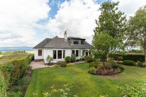 A two-story house with a sloped roof, a chimney, and a garden with various plants and a lawn. - St Michael's: Ben Ledi Views, Walk to Pubs & Explore Central Scotland (Kippen)
