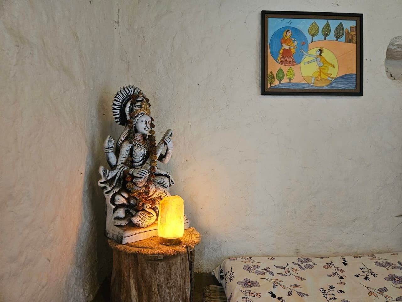 Una estatua sobre un taburete de madera con una vela encendida, un cuadro en la pared y una colcha con estampado.