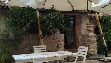 Una veranda esterna con tavolo e sedie, un ombrellone bianco e un edificio in pietra sullo sfondo.