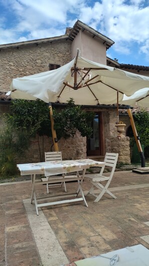 An outdoor patio with a table and chairs, a white umbrella, and a stone building in the background. - A cozy home nestled in the green hills, 4 km from the town center (Marche)