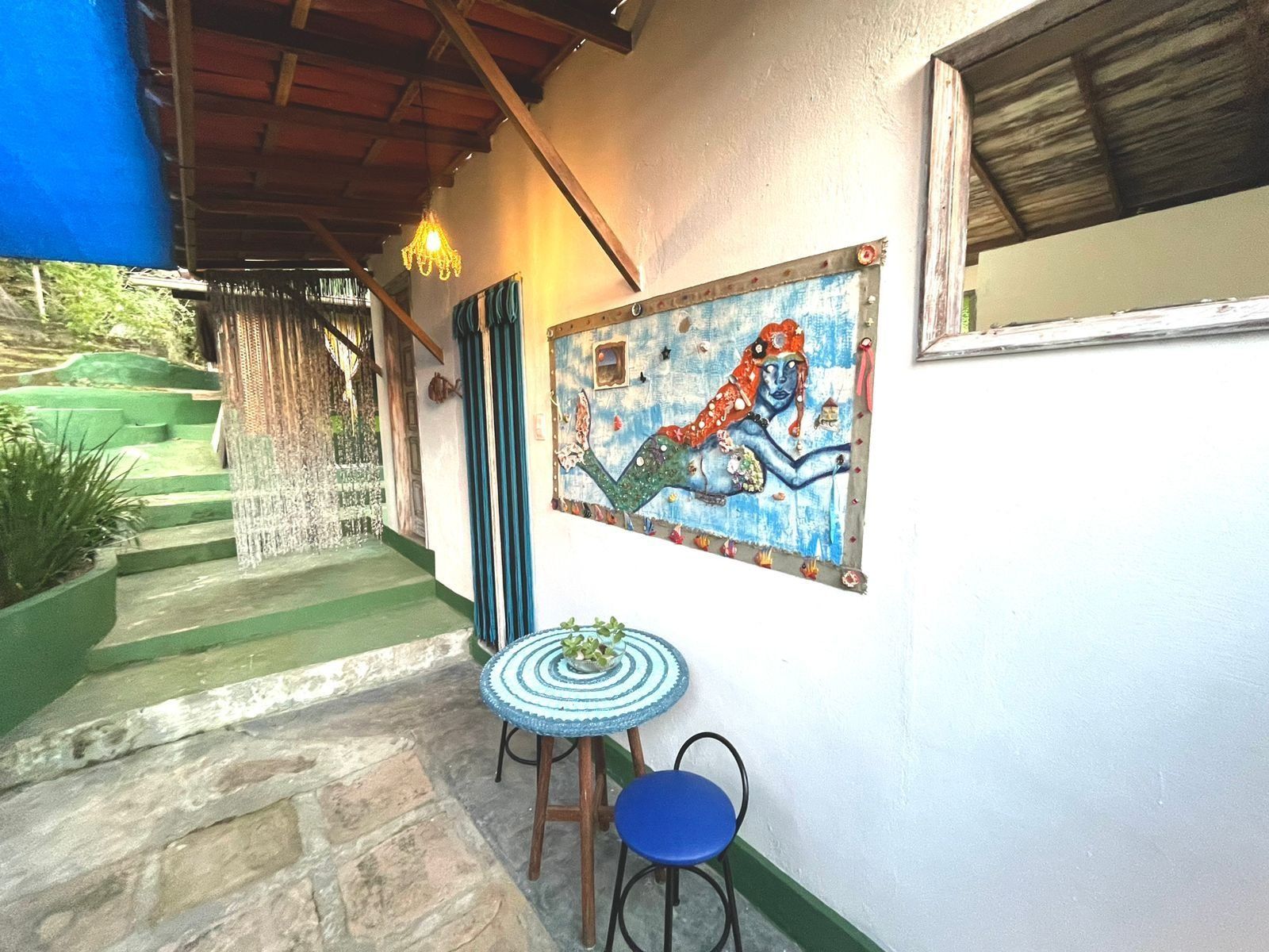 Un espacio al aire libre techado con un trabajo artístico en mosaico, una pequeña mesa redonda con una silla azul, y una escalera que lleva a una zona verde.