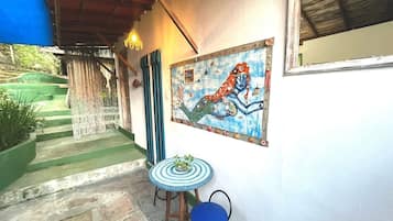 Un espacio al aire libre techado con un trabajo artístico en mosaico, una pequeña mesa redonda con una silla azul, y una escalera que lleva a una zona verde.