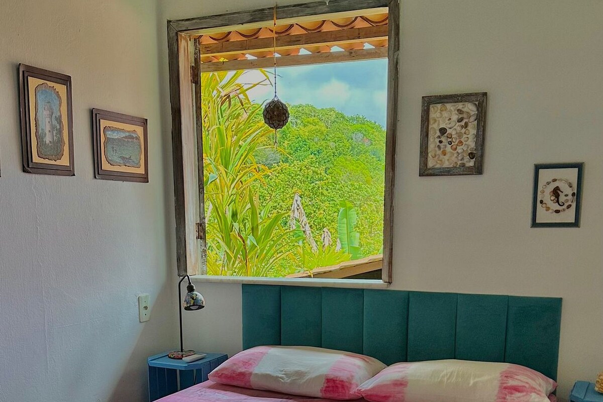 Quarto com cama, janela com vista para área verde e mesa de cabeceira com luminária.