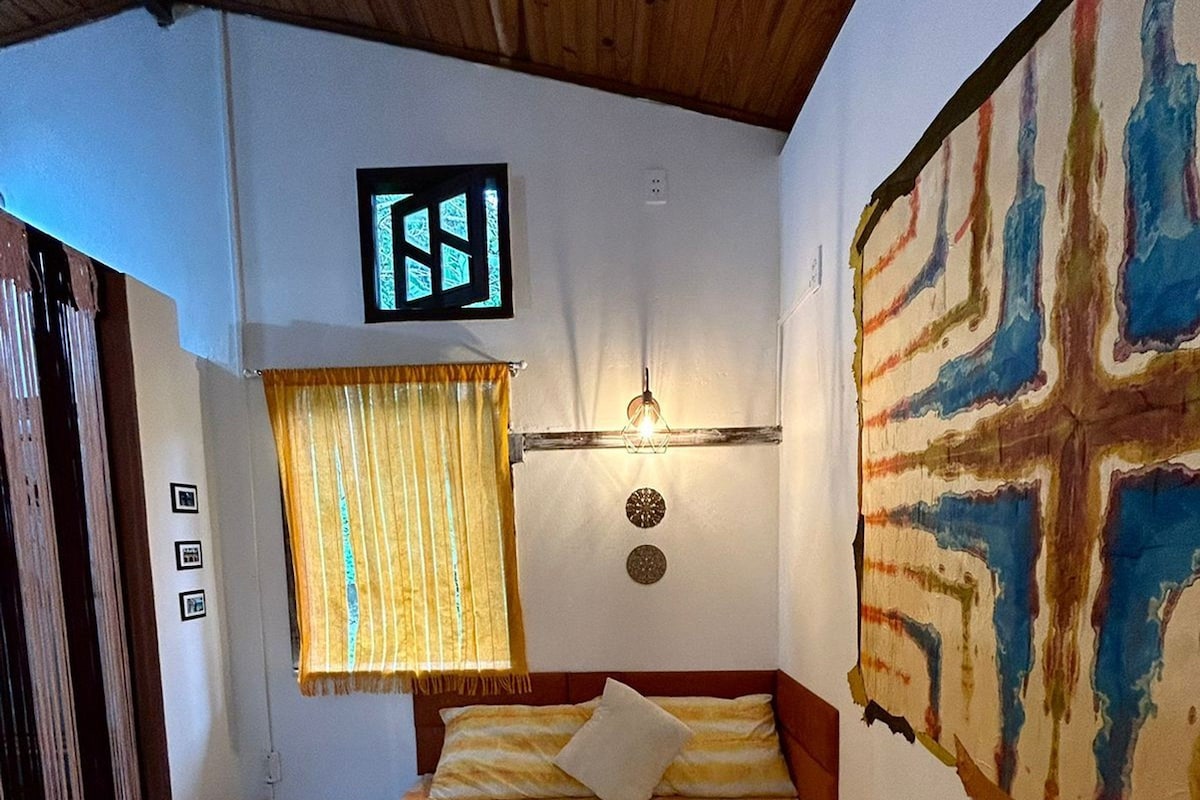 Quarto de hotel com cama, relógio de parede, janela com cortinas e uma grande pintura abstrata.