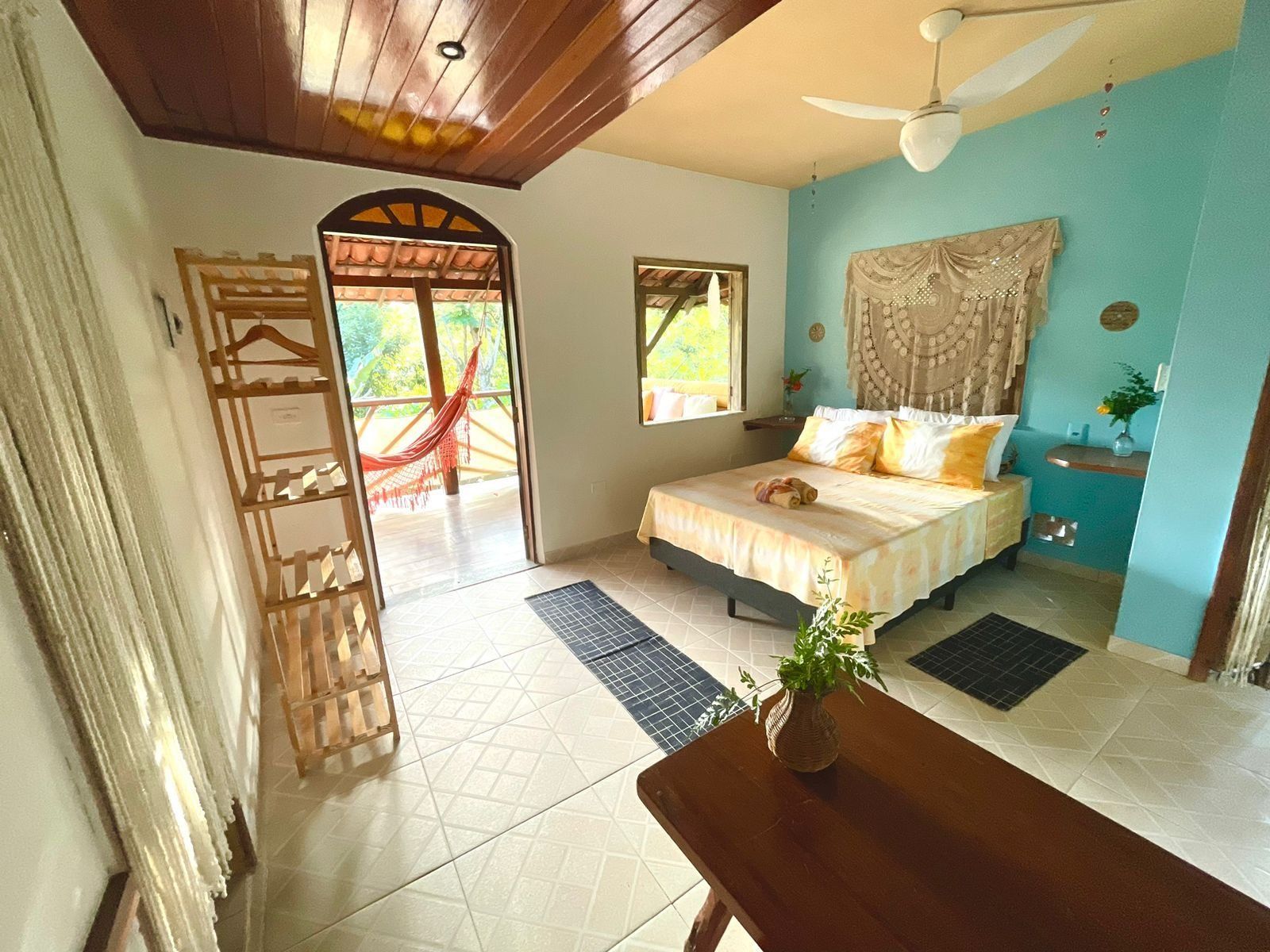 Quarto com cama, janela, ventilador de teto, rede do lado de fora e mesa de madeira com planta.