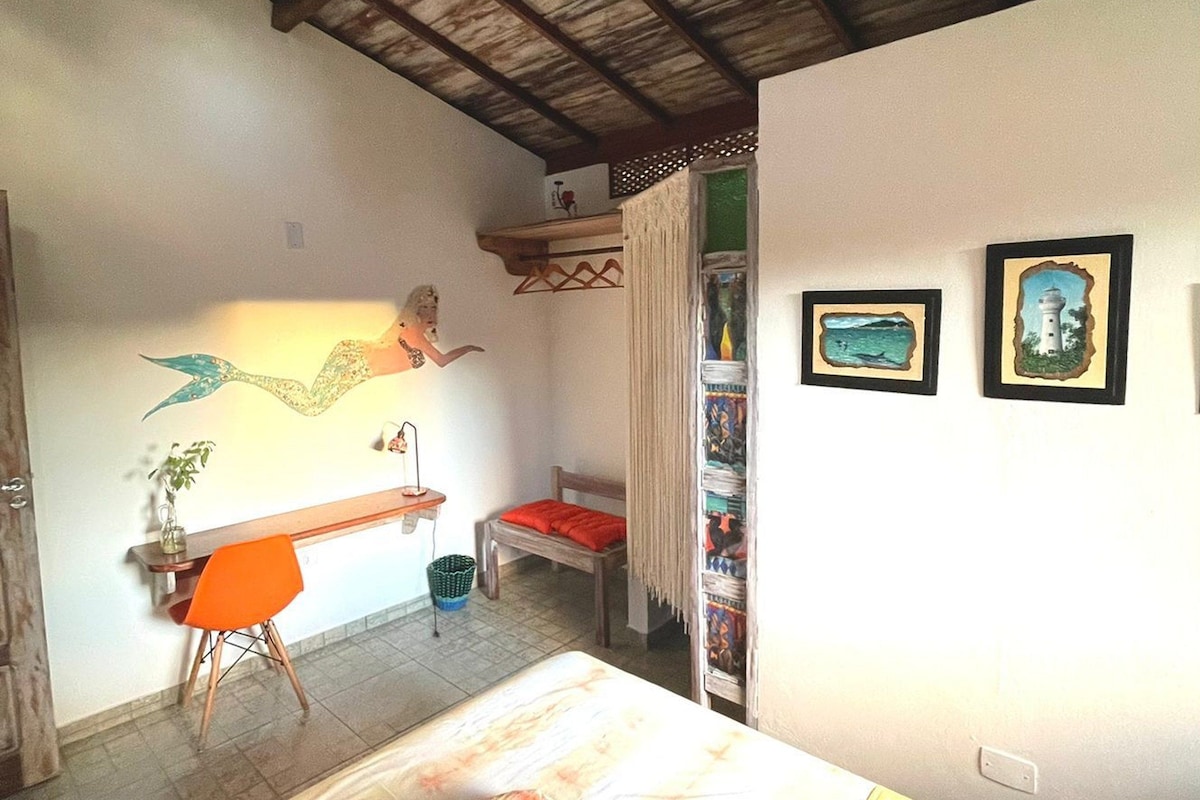 Quarto com cama, escrivaninha, cadeira, prateleira com livros e decoração de sereia na parede.