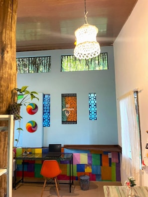 A room with a wooden ceiling, a hanging light fixture, a colorful wall with artwork, a table with a laptop, and a chair.