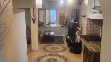 A living room with a sofa, coffee table, and a rug. There is a kitchen area with cabinets and a countertop.