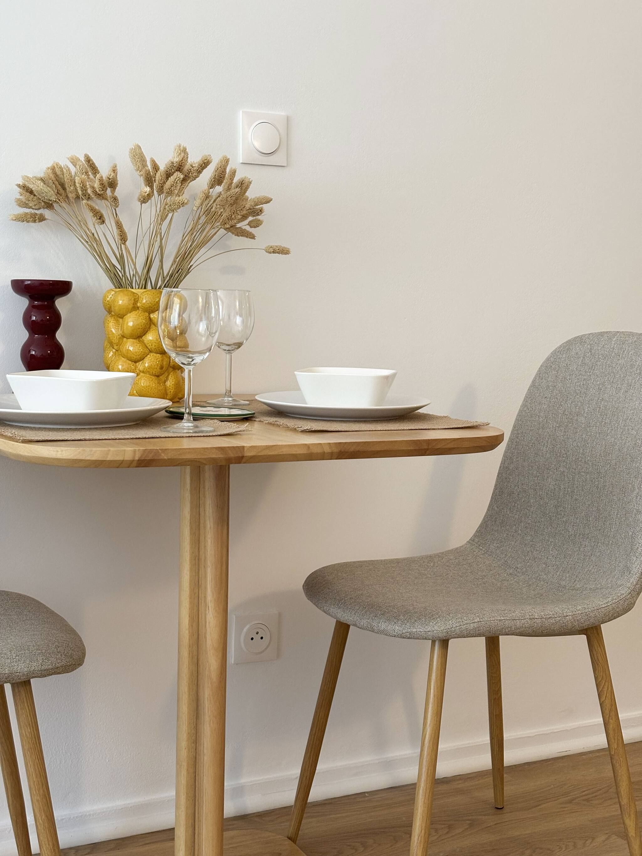 Una mesa de comedor con un jarrón de flores secas, dos copas de vino y cuencos blancos sobre una mesa de madera frente a una pared lisa.