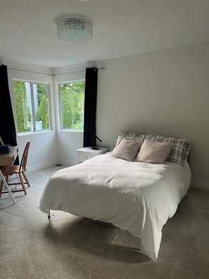 Een slaapkamer met een groot bed, een bureau en een stoel. Er zijn twee ramen met gordijnen en een plafondlamp.