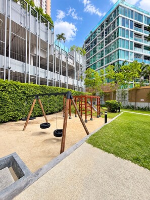 A playground with swings and a slide, surrounded by a well-maintained lawn and a building with large windows. - 10 Mins walk to Walking Street Apartment in Pattaya Jomtien (ตำบล หนองปรือ)