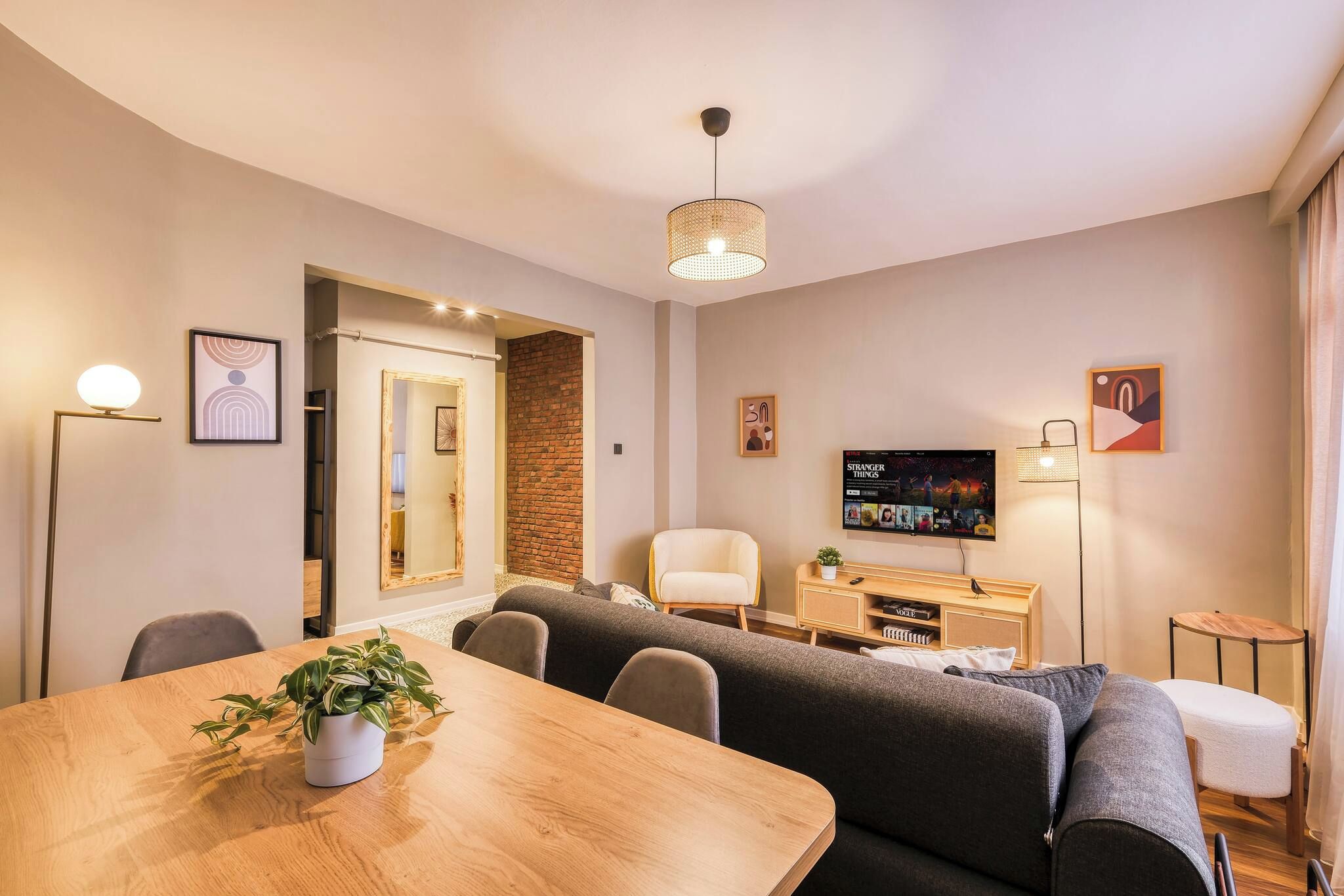 Ein modernes Wohnzimmer mit einem Holzesstisch, grauen Sofas, einem Fernseher, der 'Stranger Things' zeigt, und einer Ziegelwand als Blickfang.