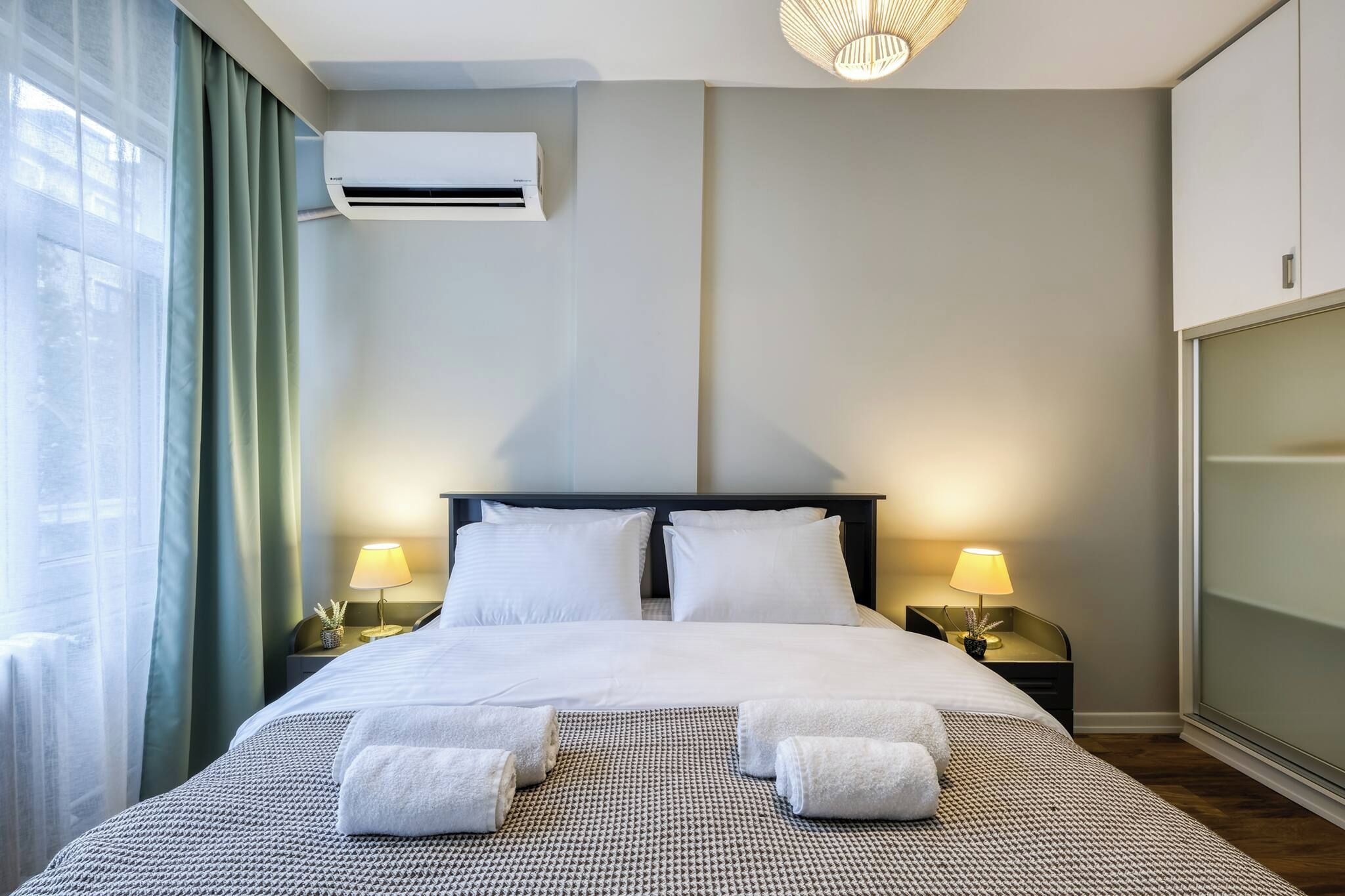 Een hotelkamer met een bed, twee bedlampen, een airconditioning en uitzicht door een raam met gordijnen.
