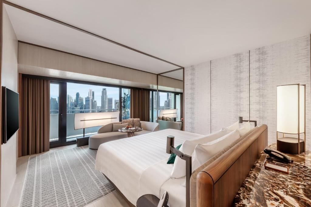 Ein modernes Hotelzimmer mit einem großen Bett, einer Sitzecke, Blick auf die Stadt und einem großen Spiegel.