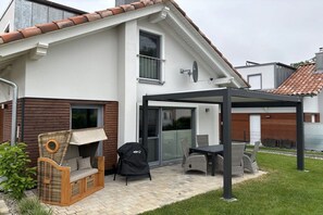 Ein Haus mit überdachter Terrasse, einem Korbsessel, einem Grill und einem Esstisch.