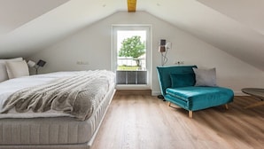 A bedroom with a bed, a teal armchair, a small table, and a window with a view of greenery. - Holiday home with 1 bedroom (Krakow am See)
