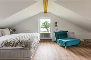 Ein Schlafzimmer mit einem Bett, einem türkisfarbenen Sessel, einem kleinen Tisch und einem Fenster mit Blick auf Grünflächen.