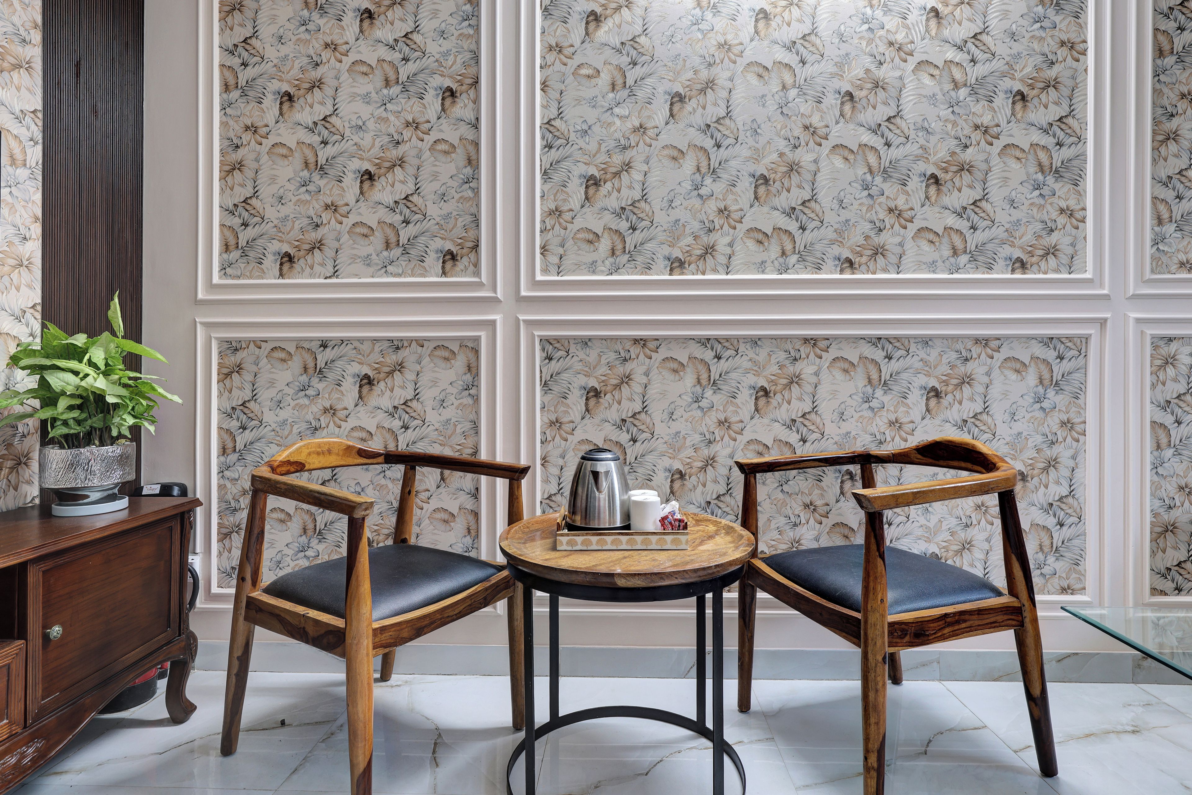 A room with a floral wallpaper, two wooden chairs with black cushions, a small round table with a kettle and cups, and a potted plant on a wooden cabinet.