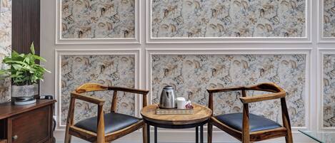 Quarto com papel de parede floral, duas cadeiras de madeira com almofadas pretas, uma pequena mesa redonda com chaleira e xícaras, e uma planta em vaso sobre um armário de madeira.