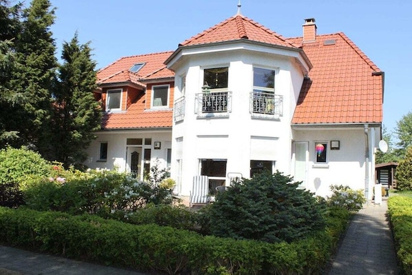 A two-story house with a red-tiled roof, a balcony with a metal railing, and a pathway leading to the entrance. - Holiday room with terrace (Zingst)