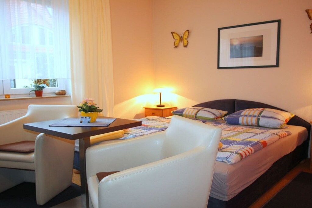 Une chambre d’hôtel comprenant un lit, un bureau, une chaise, une lampe, une fenêtre avec des rideaux, une plante et un élément décoratif en forme de papillon.