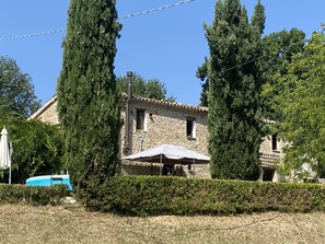 Ein Gebäude aus Stein mit einem Ziegeldach, umgeben von hohen Bäumen und einer Hecke.