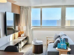 A modern hotel room with a large TV, a sofa, a coffee table with water bottles, and a view of the ocean. - 40F-High Floor-Stunning Ocean View-1 Bedroom Condo-Waikiki Beach (Honolulu)