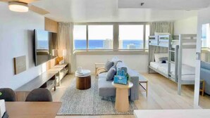 A modern hotel room with a large window, a flat-screen TV, a sofa, a dining area, and bunk beds. - 40F-High Floor-Stunning Ocean View-1 Bedroom Condo-Waikiki Beach (Honolulu)