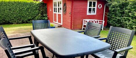 An outdoor patio with a table and chairs, a red building in the background, and a hedge.