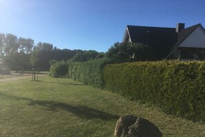 A grassy area with a hedge, a stone, and a building with a dark roof. - Holiday apartment MEERzeit (Rerik)