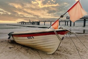 Ein Boot mit der Aufschrift 'KOS 6' an einem sandigen Strand, im Hintergrund ein Steg.
