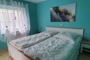 A bedroom with a bed, a window with sheer curtains, a nightstand, and a wall-mounted picture. - Holiday apartment with 1 bedroom (Sassnitz)
