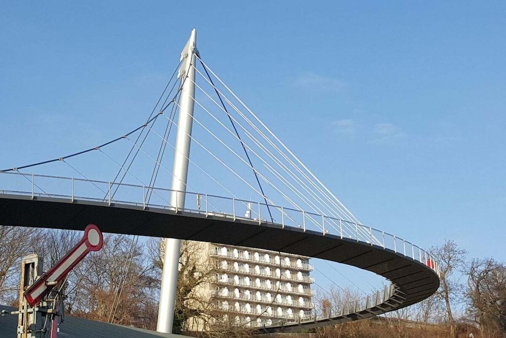 A pedestrian bridge with a curved design, a tall central support, and a building with multiple balconies in the background.