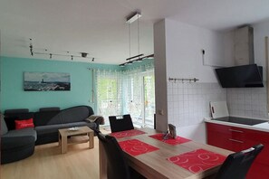 A modern living room with a sofa, a dining table with chairs, a kitchen area with a TV, and a large window with curtains. - Holiday apartment with 1 bedroom (Sassnitz)
