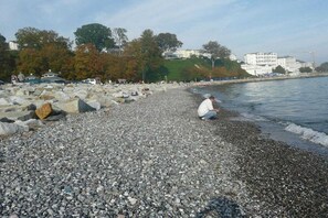 A pebbled beach with a person sitting on the shore, large rocks, and buildings in the background. - Holiday apartment with 1 bedroom (Sassnitz)