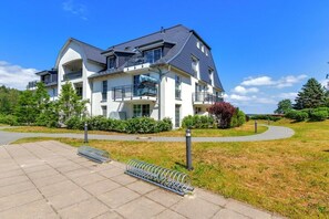 A multi-story residential building with balconies, a paved driveway, and a grassy area with a metal bike rack. - Holiday apartment in Benz with swimming pool (Benz)