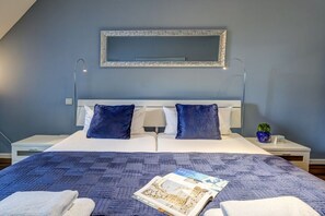 Un letto rifatto con una coperta fantasia, due cuscini e una rivista sopra. Un comodino con una lampada e una piccola pianta accanto al letto.
